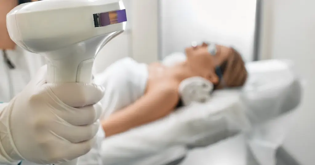 Gloved hand holds a white laser device for Lumecca IPL in Owensboro, KY, as a woman in goggles lies on a treatment table.